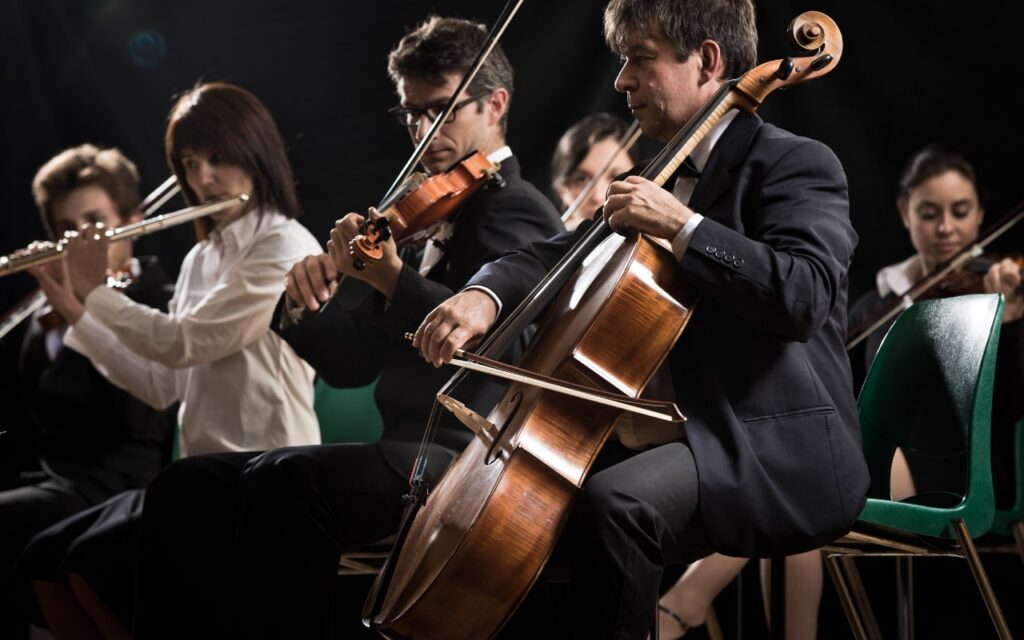 A group of musicians in formal attire perform on stage, playing cello, violin, flute, and viola. They are seated in a row, focused on their instruments, with a dark background behind them.