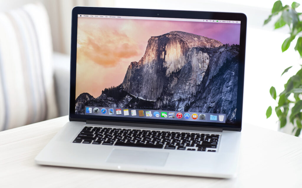 A silver MacBook laptop with a Yosemite National Park wallpaper displayed on its screen sits open on a light-colored table, with a blurred plant and striped cushions in the background.