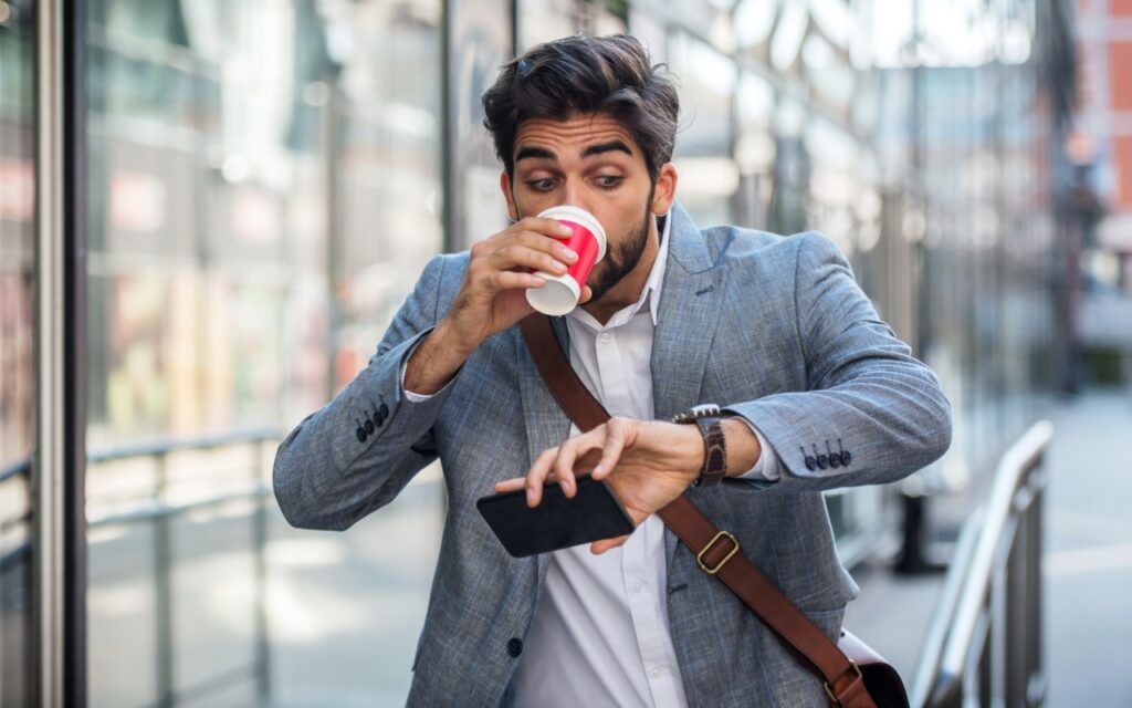A man in a suit jacket drinks coffee from a red cup while hurriedly looking at his watch and holding a smartphone. He appears to be in a rush, standing outdoors in an urban setting.