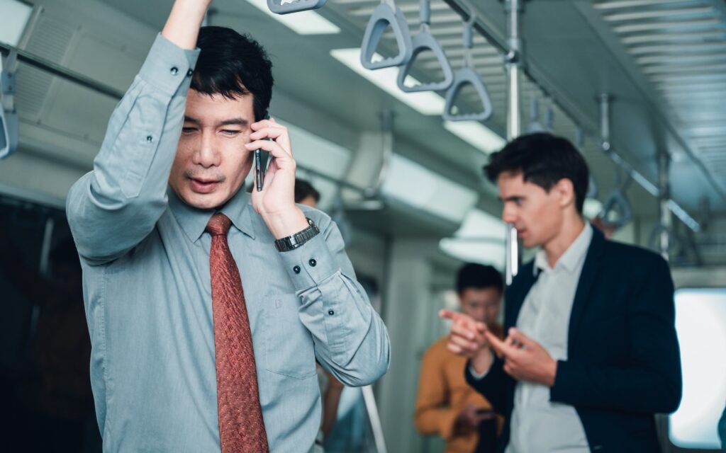 A man in business attire holds onto a subway handrail and talks on his phone, while two other men in the background have a conversation inside the train.