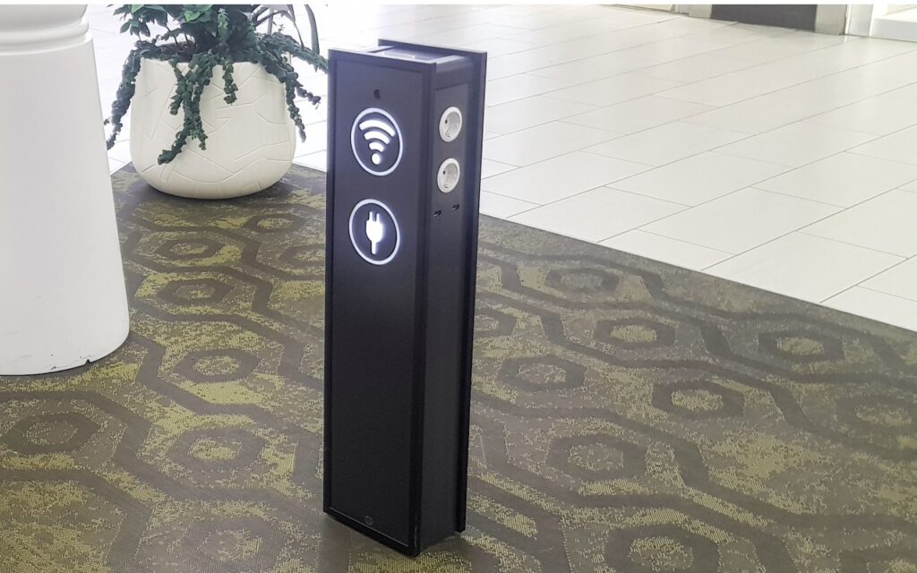 A tall black charging station with illuminated WiFi and plug icons stands on a patterned carpet in a public indoor space, next to a large white planter with green plants.