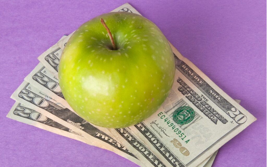 A green apple resting on top of several U.S. twenty dollar bills, all placed on a purple surface.