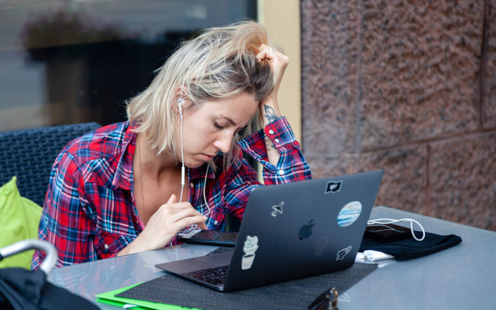 A person with blonde hair in a red plaid shirt sits at a table, looking stressed while working on a laptop. They are wearing earphones and resting their head on one hand.
