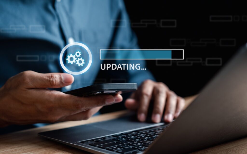 A person holds a smartphone and uses a laptop, with a digital overlay showing gears and a progress bar labeled UPDATING... indicating a software or system update in progress.