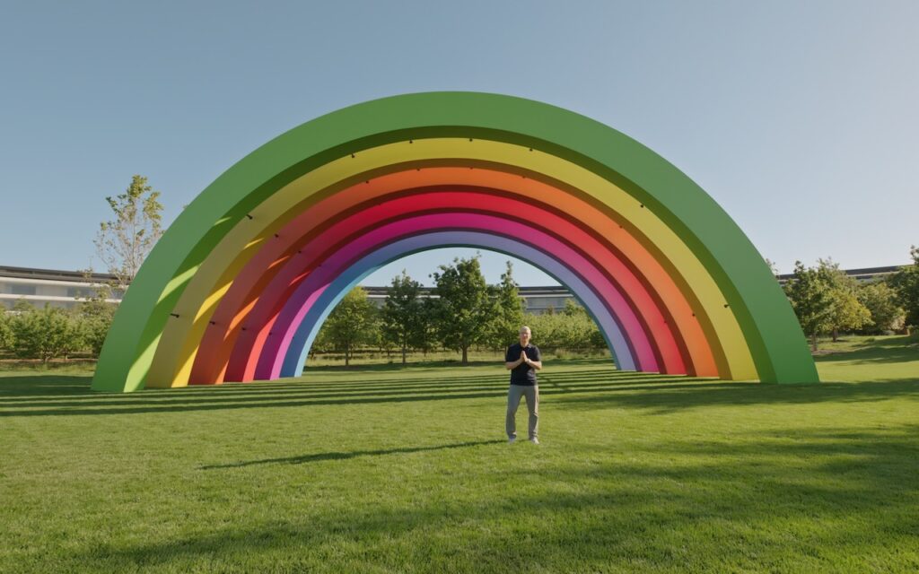 A person stands on grass in front of a large, colorful rainbow-shaped arch sculpture in a park with trees and clear blue sky in the background.
