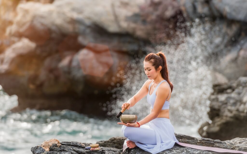 A woman in light workout clothes sits cross-legged on rocks by the sea, playing a singing bowl with a mallet. Waves crash against the rocks in the background, creating a serene, meditative atmosphere.