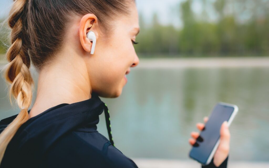 A woman with a braided ponytail, wearing wireless earbuds and a black jacket, holds a smartphone while standing outdoors near a body of water with greenery in the background.
