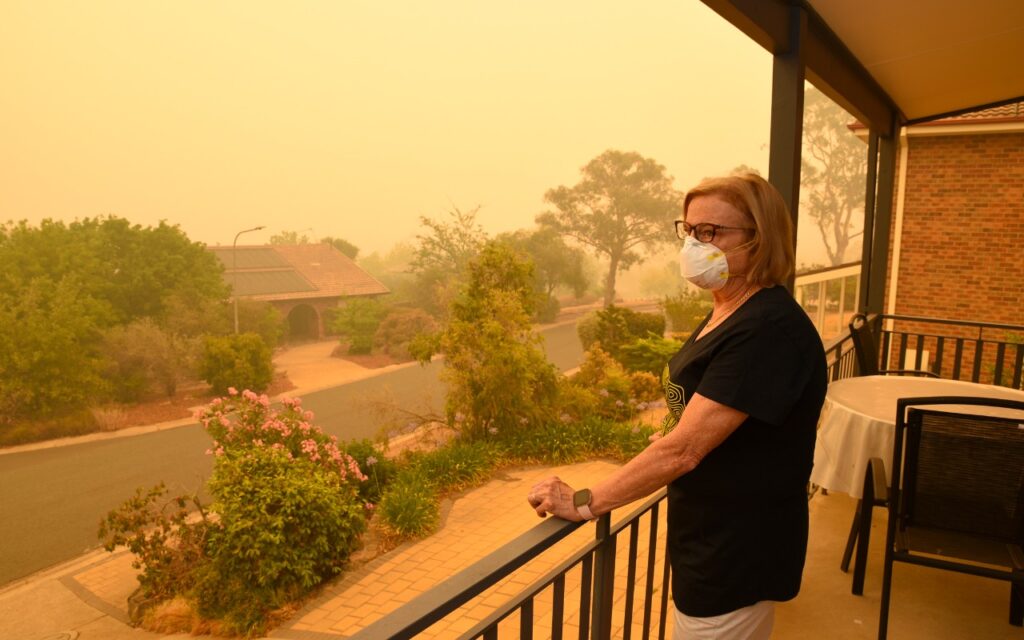 A woman wearing a mask stands on a porch, looking at a street and garden outside. The air is thick with smoke, and visibility is low due to what appears to be a wildfire or heavy pollution.