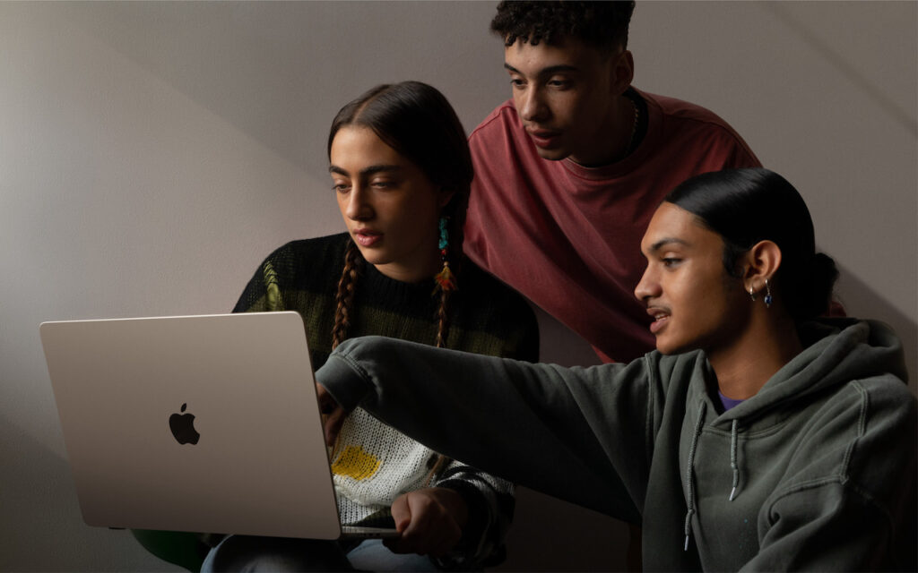 Three young people sit closely together, looking at a laptop screen. One person points at the screen while the others watch attentively, appearing to collaborate or study. The setting is indoors with neutral lighting.