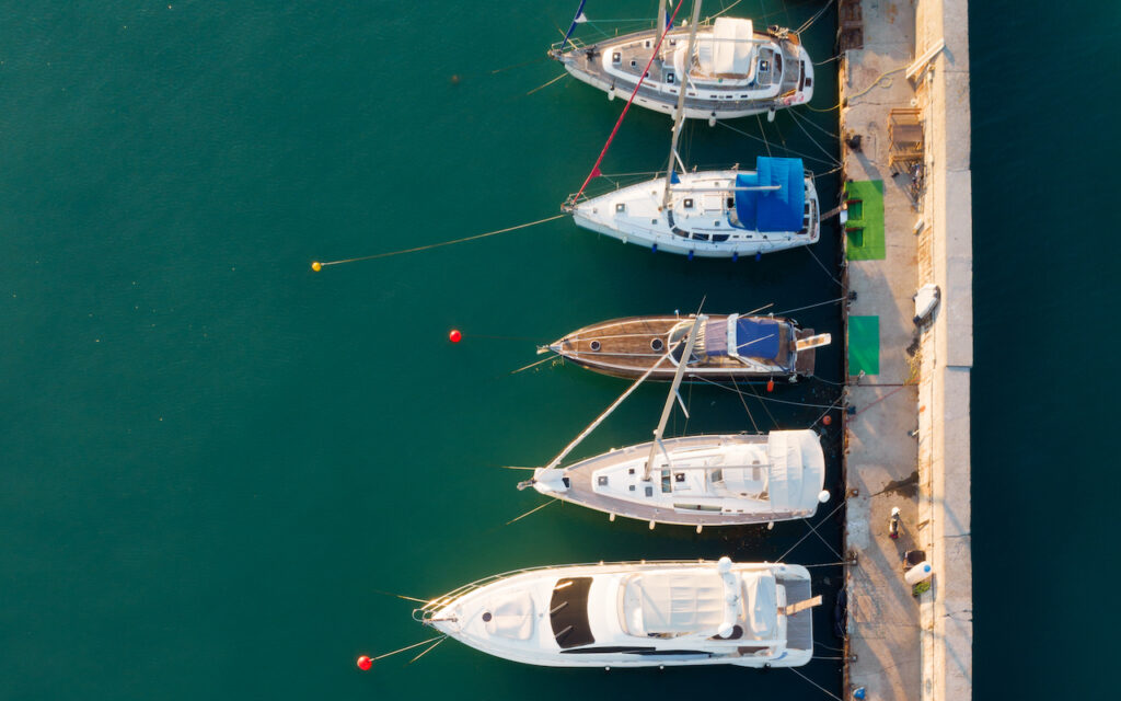 Aerial view of four yachts docked side by side along a pier in clear turquoise water, with ropes and small green mats visible on the pier.