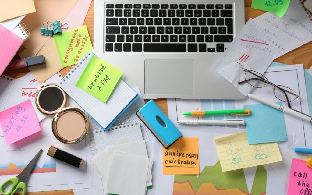 A cluttered desk with a laptop, notepads, sticky notes with reminders, makeup items, eyeglasses, papers, charts, and stationery scattered around, suggesting a busy or disorganized workspace.