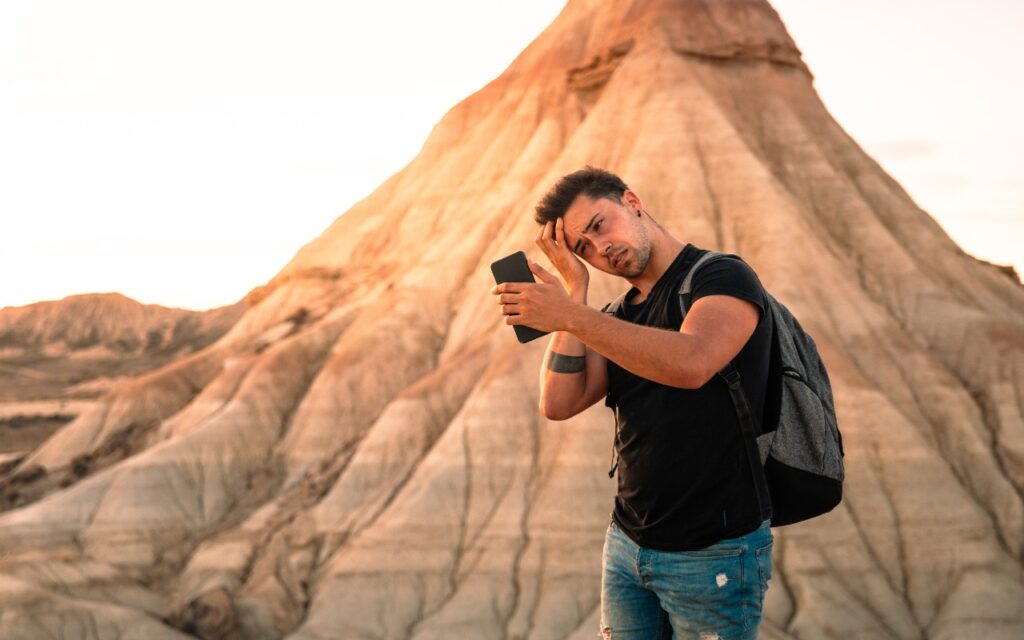 A man with a backpack stands in front of a large rocky hill, holding a phone and touching his hair, as if taking a selfie. The landscape is arid with soft sunlight highlighting the scene.