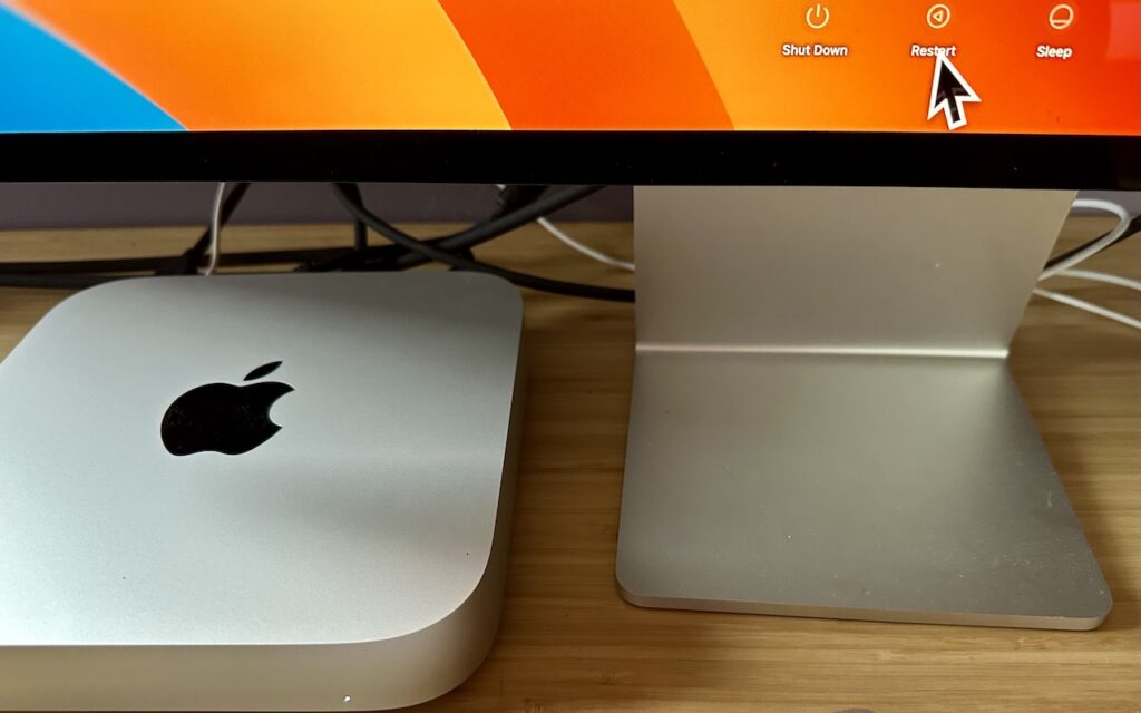 A close-up of a Mac Mini next to an iMac stand on a wooden desk. On the monitor, a pointer hovers over the Restart option in the macOS shutdown menu. Cables are visible behind the devices.