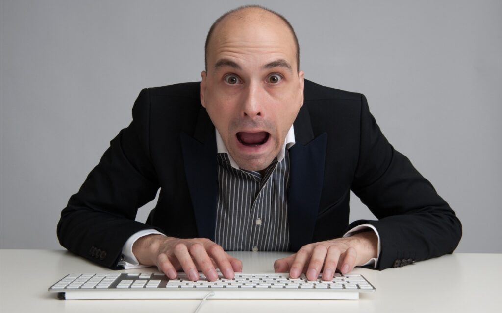 A man in a suit with a surprised or shocked expression types on a keyboard at a desk, leaning forward with wide eyes and mouth open.