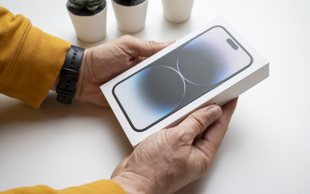 A person wearing a yellow sleeve and a black watch holds a boxed smartphone, likely an iPhone, on a white table with three small potted plants in the background.