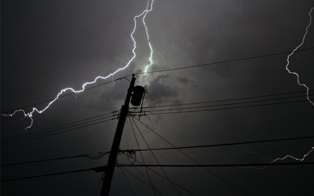A bolt of lightning flashes across a dark, cloudy night sky above a silhouetted utility pole and power lines.