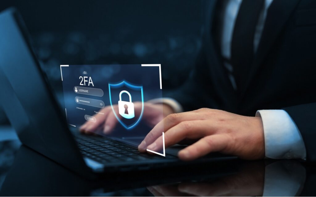 Person in a suit typing on a laptop with a digital overlay showing a shield, padlock, and “2FA” (two-factor authentication) login screen, representing cybersecurity and secure access.