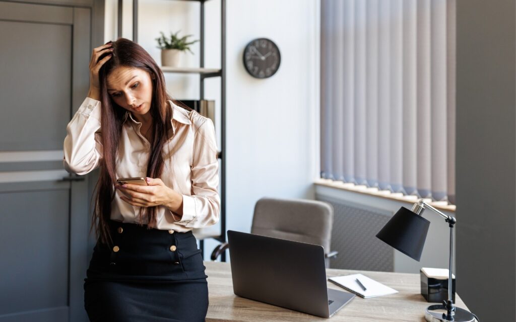 A woman with long brown hair stands in an office, looking worried as she holds her phone and touches her head. She leans on a desk with a laptop, documents, and a lamp. A clock and shelves are in the background.