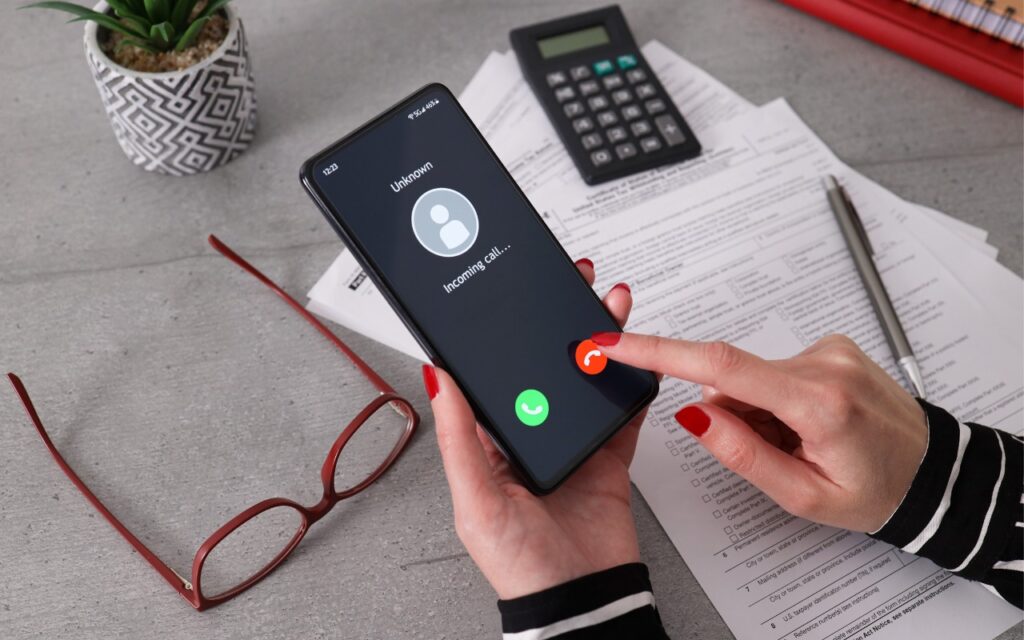 A person with red painted nails holds a smartphone displaying an incoming call from Unknown. The phone is on top of paperwork with a calculator, eyeglasses, pen, and potted plant nearby on a gray desk.