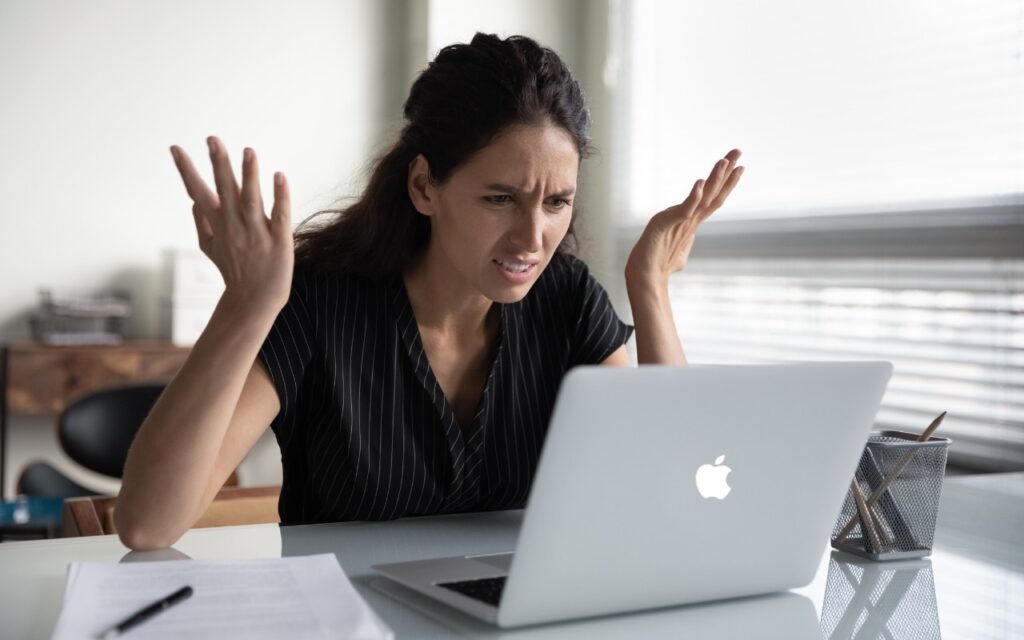 A woman sits at a desk looking frustrated with her laptop, raising her hands in confusion. Papers and a pen are on the table beside her, and light streams through window blinds in the background.