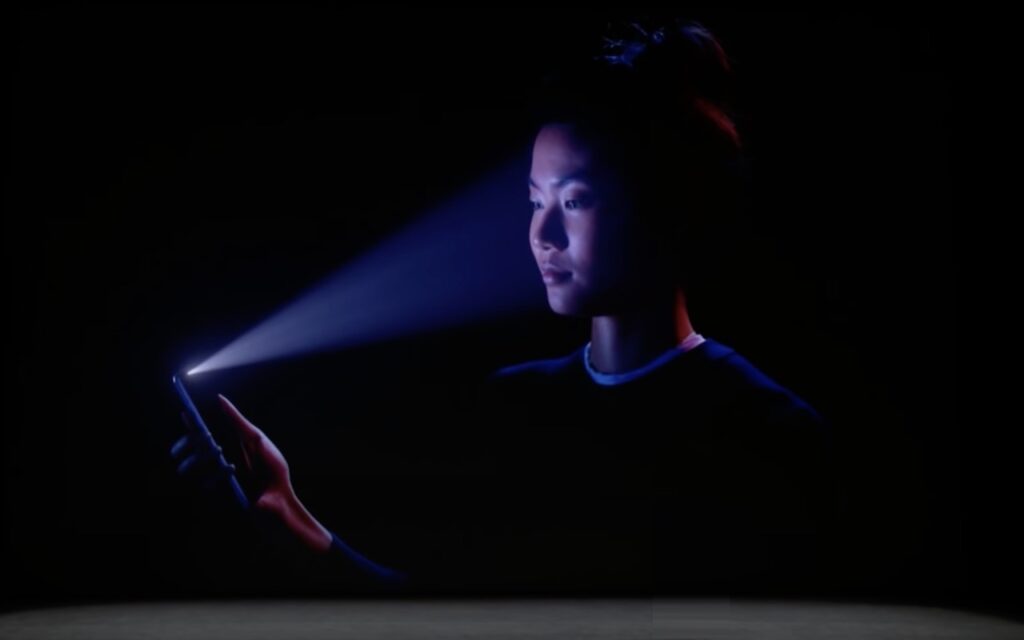 A woman in a dark setting holds a smartphone in front of her face, with blue light projecting from the phone onto her face, illustrating facial recognition technology.