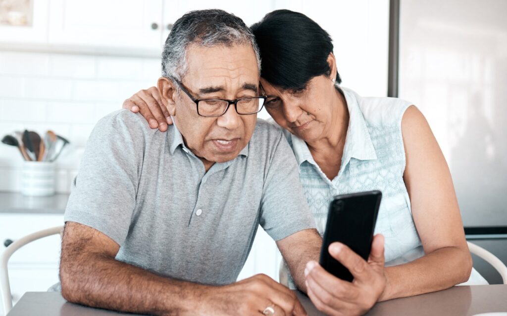 An older couple sits closely together at a table, looking concerned as they read something on a smartphone. The man wears glasses and a gray polo, while the woman leans her head on his shoulder, showing support.