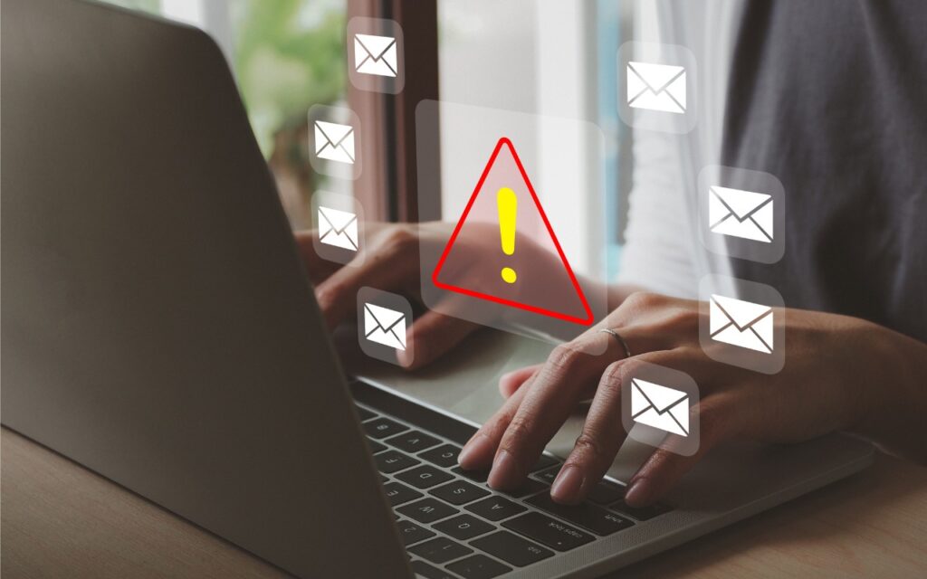 A person types on a laptop keyboard. Multiple email icons and a large red warning triangle with an exclamation mark appear digitally, suggesting an email security or phishing alert.