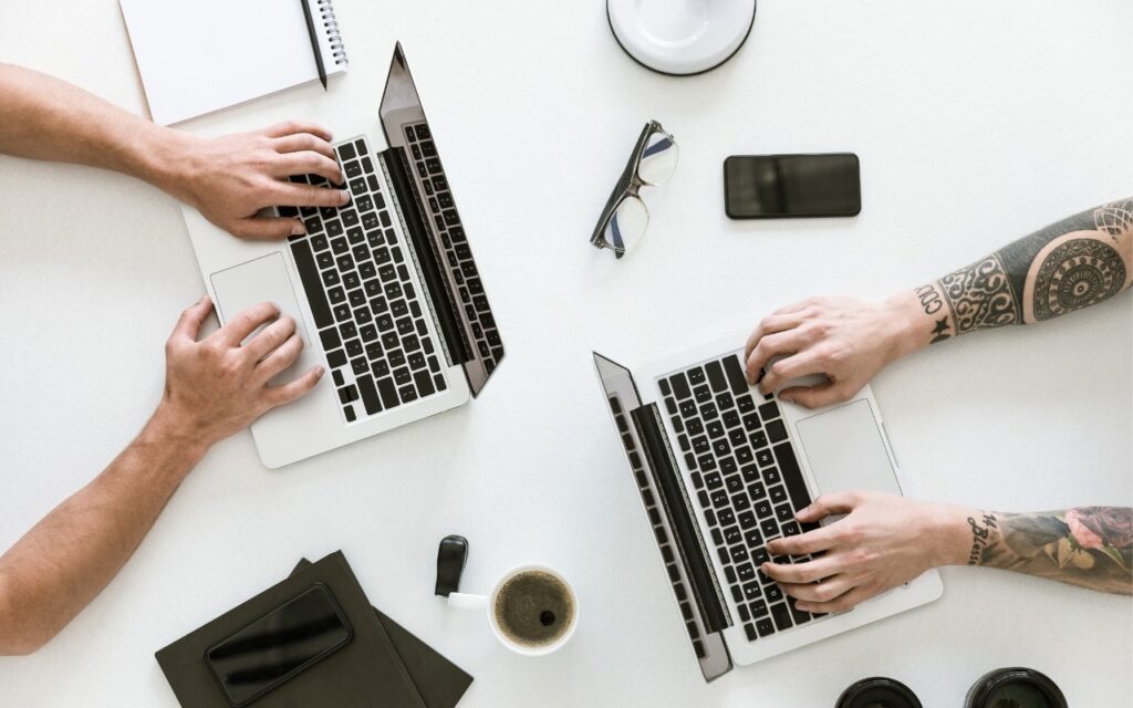 Two people working on laptops at a white desk, surrounded by notebooks, a smartphone, glasses, a cup of coffee, and a camera lens. One person has tattoos on their right arm.