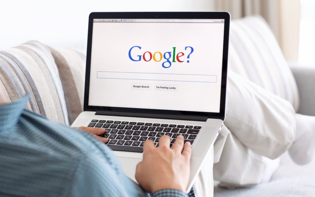 A person sits on a couch with striped pillows, using a laptop on their lap to view the Google homepage, featuring a question mark—perhaps concerned by the privacy or results of Google Search? Try these other search engines.