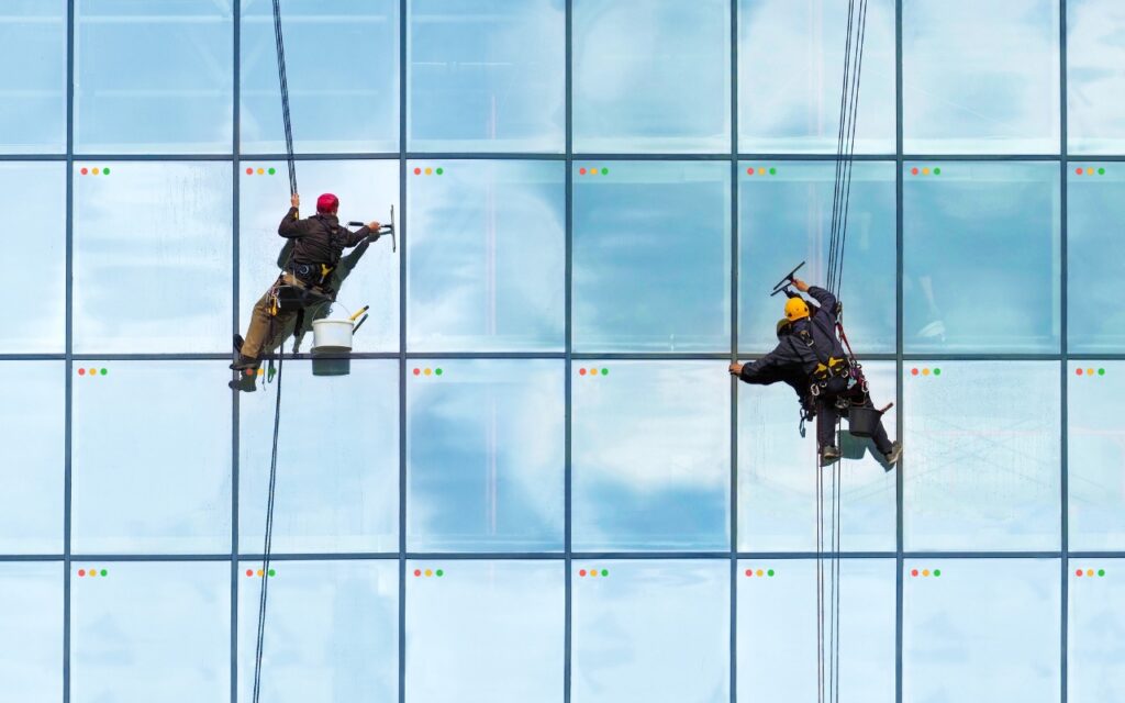 Two workers wearing safety gear are suspended by ropes, cleaning the windows of a tall glass building—reminding you that if you have too many windows open, close them all quickly with these tricks for a clearer view.