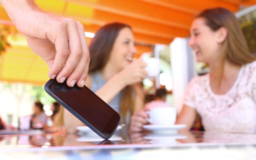 A hand stealthily takes a smartphone from a table while two women sit in the background, smiling and drinking coffee, unaware of the theft—use iOS 17.3’s Stolen Device Protection to reduce harm from iPhone passcode thefts.