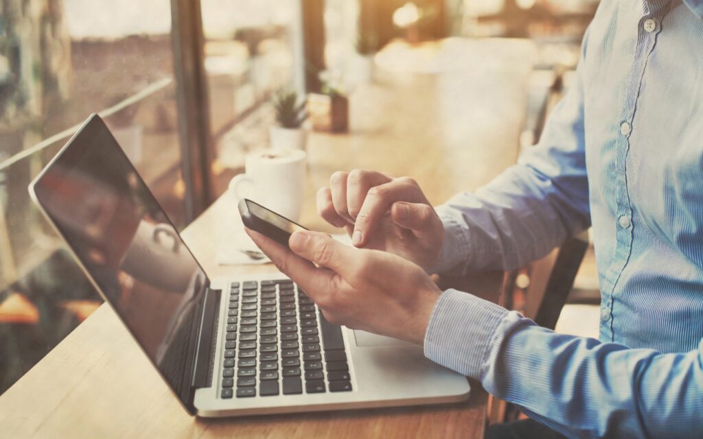 A person in a blue shirt uses a smartphone while sitting at a table with an open laptop, exploring how to sync your text messages across all your Apple devices in a bright café with a window and cup in the background.