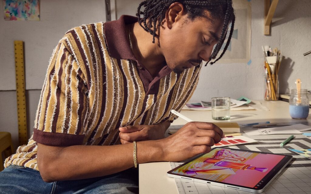 A person with braided hair uses an Apple Pencil Pro to draw colorful digital art on an iPad Pro at a desk, surrounded by art supplies, paper swatches, and a glass of water.
