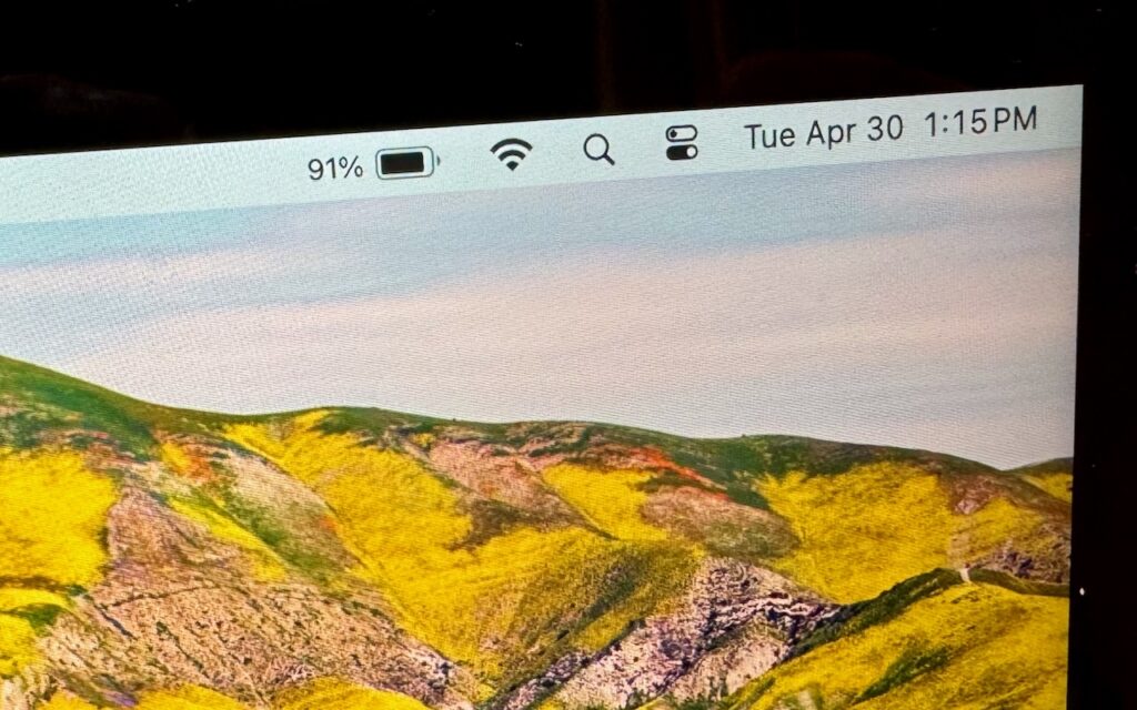 A close-up of a computer screen shows 91% battery, Wi-Fi, and control center icons—demonstrating how to display the battery percentage in your Mac’s menu bar. The background displays hills with green and yellow vegetation.