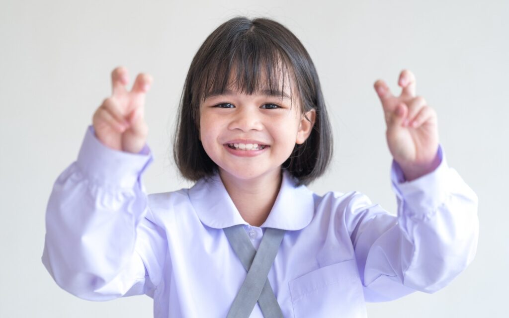 Smiling young girl in a white shirt making air quotes with both hands, standing against a plain light background—perfect for illustrating "Where Can You Control Automatic Smart Quotes and Dashes in macOS?" in a fun, relatable way.