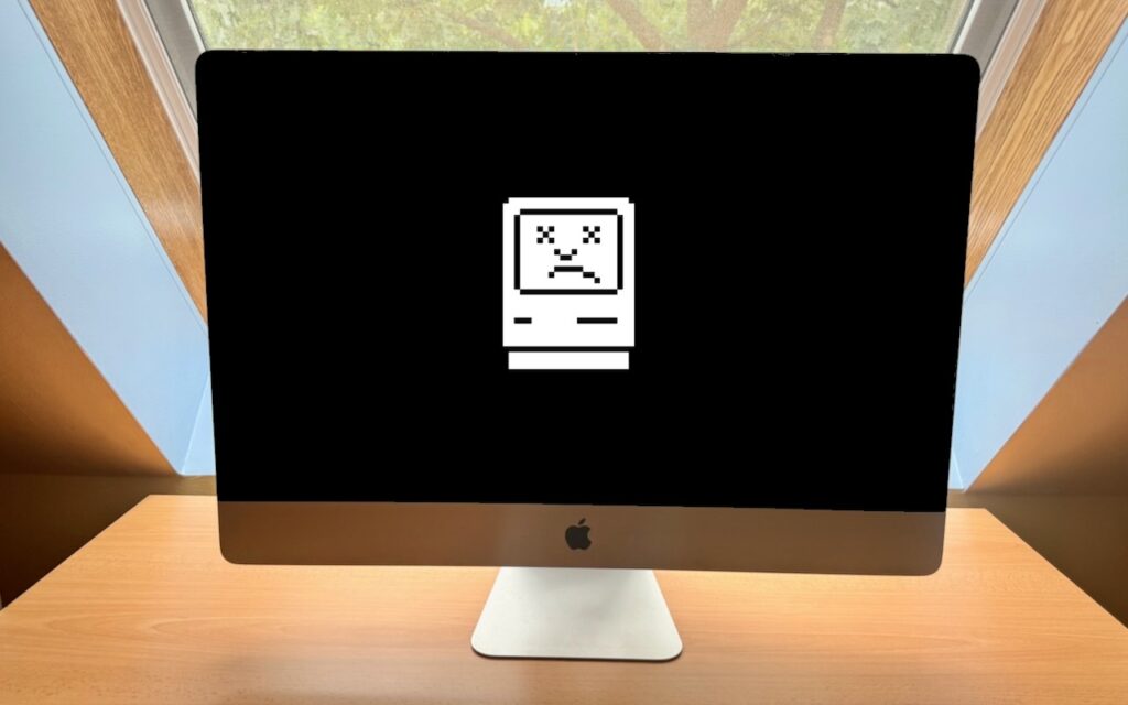A desktop iMac displays a black screen with a white, pixelated sad Mac icon, indicating a system error—perhaps reflecting the news: Sorry, Apple Isn’t Going to Update the 27-inch iMac with Apple Silicon. The computer sits on a wooden desk near a window.