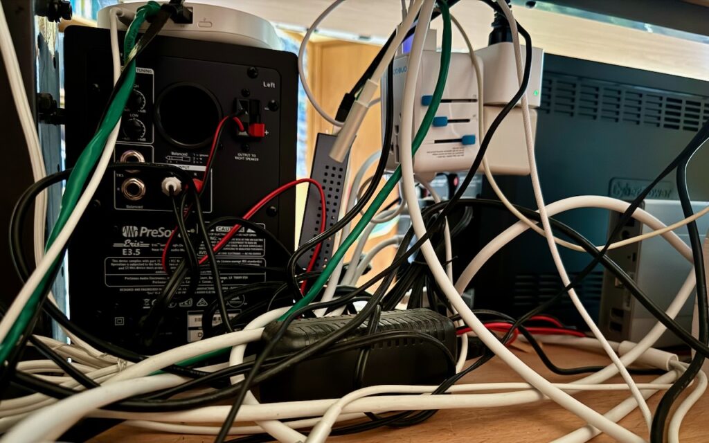 A cluttered workspace with numerous tangled cables connected to electronic equipment, including a PreSonus speaker, a power strip, a monitor, and various adapters on a wooden table. Tame the Tangle! Quick Cable Management Tips can help organize your setup.