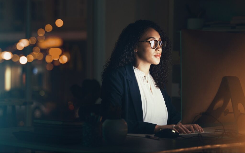 A woman wearing glasses and business attire works at a computer in a dimly lit office at night, illuminated by the screen and city lights outside. Working late on your Mac? Turn on Night Shift to help your sleep.