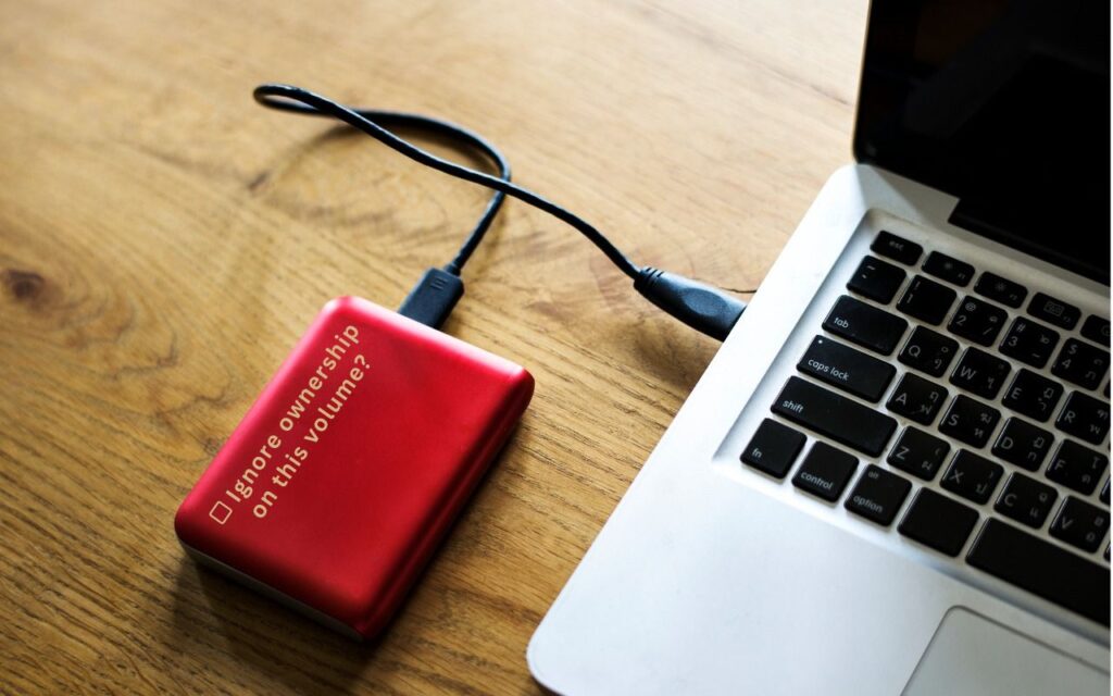 A red external hard drive with the text Ignore ownership on this volume? is connected to a silver laptop on a wooden surface, prompting the question: When Should You “Ignore Ownership” on an External Drive?.