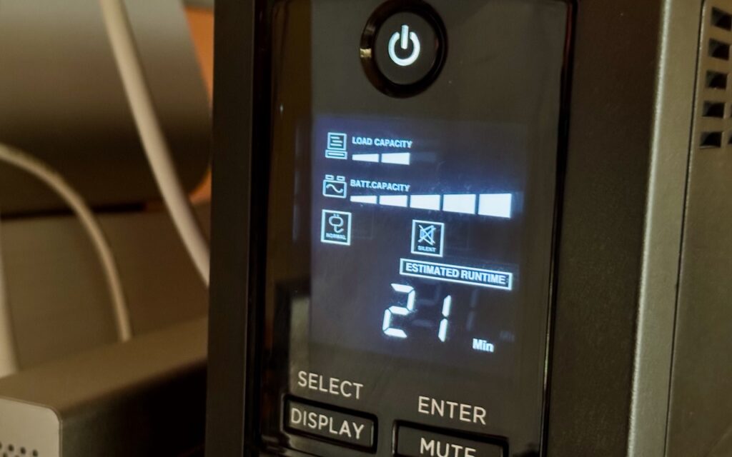 Close-up of a UPS display showing full battery and load capacity, plus an estimated 21-minute runtime. Buttons labeled Select, Display, Enter, and Mute are visible. Energy Saver can shut down your UPS-equipped Mac in a power outage.
