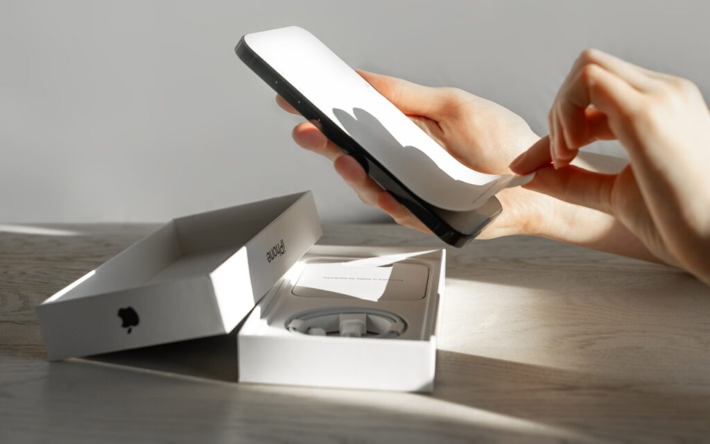A person’s hands are unboxing a new smartphone, peeling protective film off the device—what you need to know before switching to a new iPhone is displayed by the open white Apple box and accessories on a light wood surface in bright natural light.