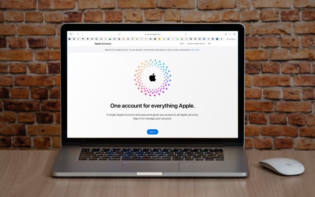 A laptop on a wooden desk displays the Apple Account login page with the message “One account for everything Apple,” highlighting that Apple ID is renamed to Apple Account in the latest operating system releases. A wireless mouse sits nearby against a brick wall backdrop.