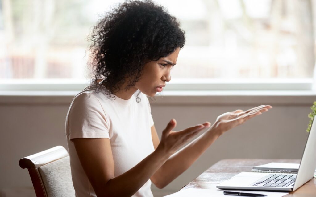 A woman with curly hair sits at a table in front of a laptop, looking frustrated and confused—perhaps struggling to figure out how to minimize downtime and lost work from tech failures. Bright window light shines behind her.