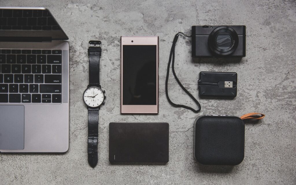 A Few of Our Favorite Things: Tech Edition—a flat lay of electronic gadgets on a gray surface, featuring a laptop, wristwatch, smartphone, camera, USB drive, external hard drive, and a small portable speaker.
