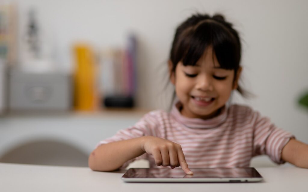 A young girl wearing a striped shirt smiles while using her finger to interact with a tablet, where Guided Access securely allows her to use an app on her iPad in a brightly lit room.