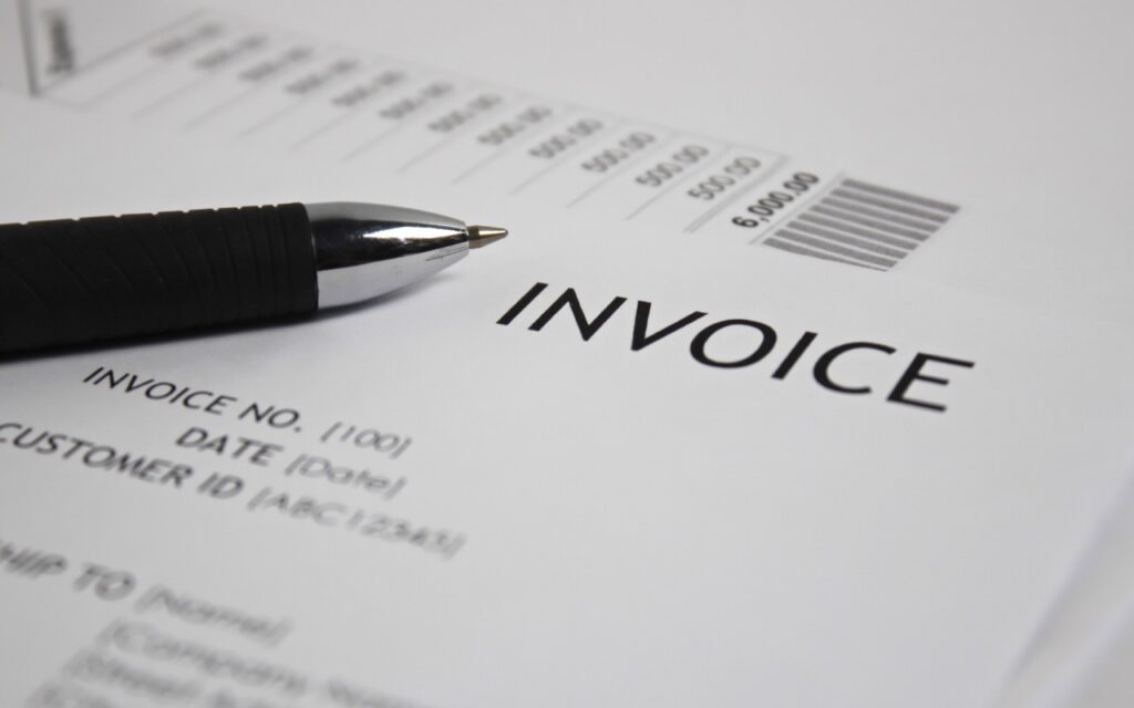 A close-up of a black pen resting on a printed invoice, with the word INVOICE clearly visible and a list of amounts and a barcode in the background. Watch out for PayPal invoice phishing scams.