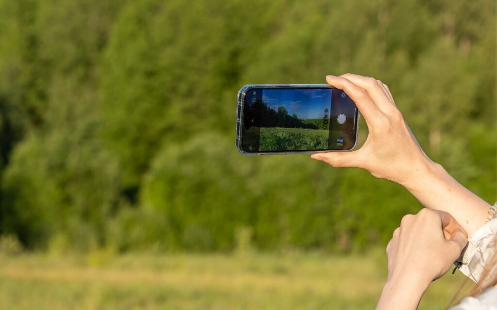 A person holds a smartphone horizontally, taking a photo of a green, sunlit forest—perhaps practicing how to train yourself to use the iPhone 16’s new camera control button. Only the person's hands are visible as they capture the bright, outdoor scene.