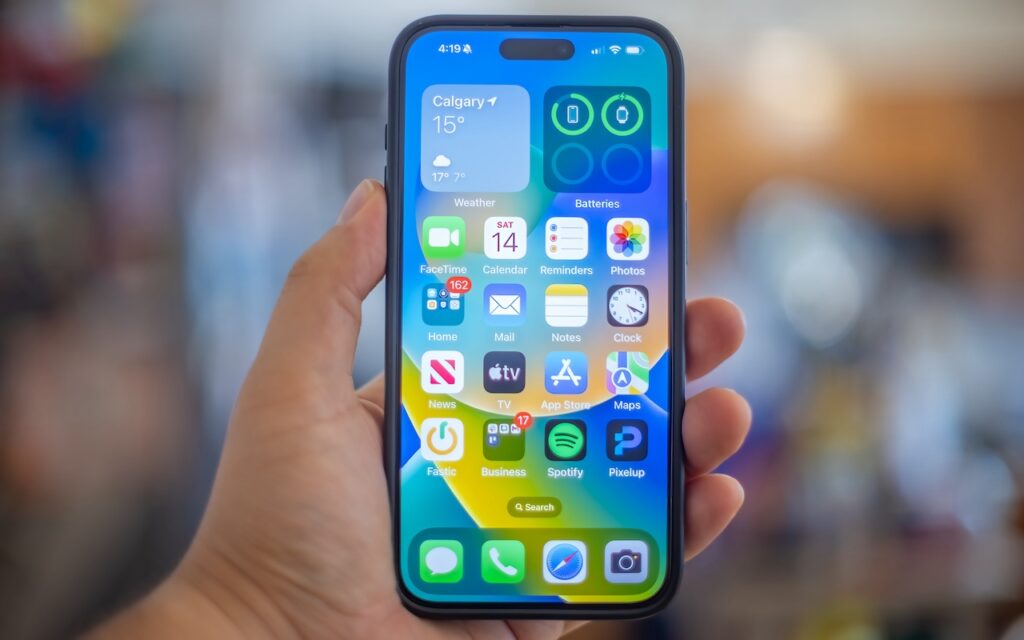 A hand holds an iPhone displaying the home screen with various app icons and a weather widget for Calgary. In iOS 18, you can quickly turn home screen app icons into widgets for enhanced customization and convenience.