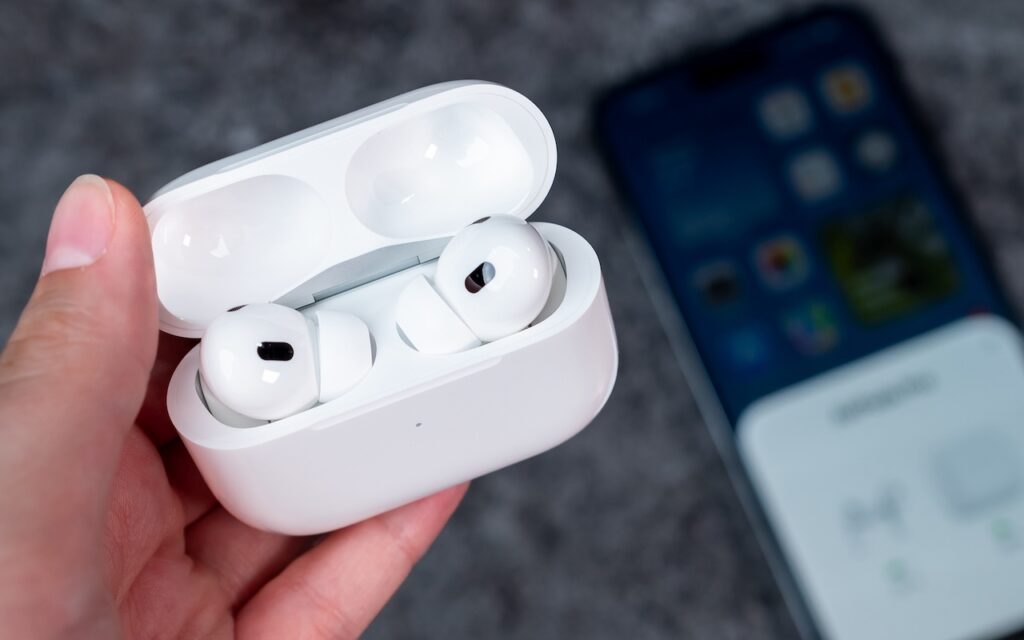 A hand holds an open white charging case with wireless earbuds inside, while a blurred smartphone lies on a gray surface in the background—highlighting how you can improve your hearing with the AirPods Pro 2 and iOS 18.