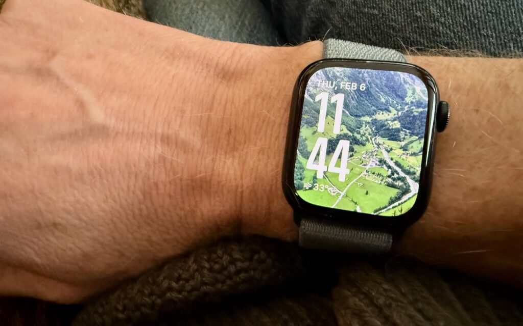 A smartwatch on a person’s wrist displays 11:44 and Thursday, February 6. The watch face background shows an aerial view of green fields and trees—For Moments of Joy, use watchOS 11’s Photos Watch Face to personalize your display.